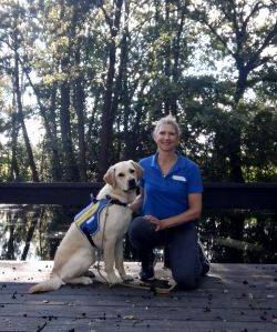 Marlies is knelling on a wooden path with the forest behind her. A yellow labrador in blue assistance dog jacket is sitting next to her.
